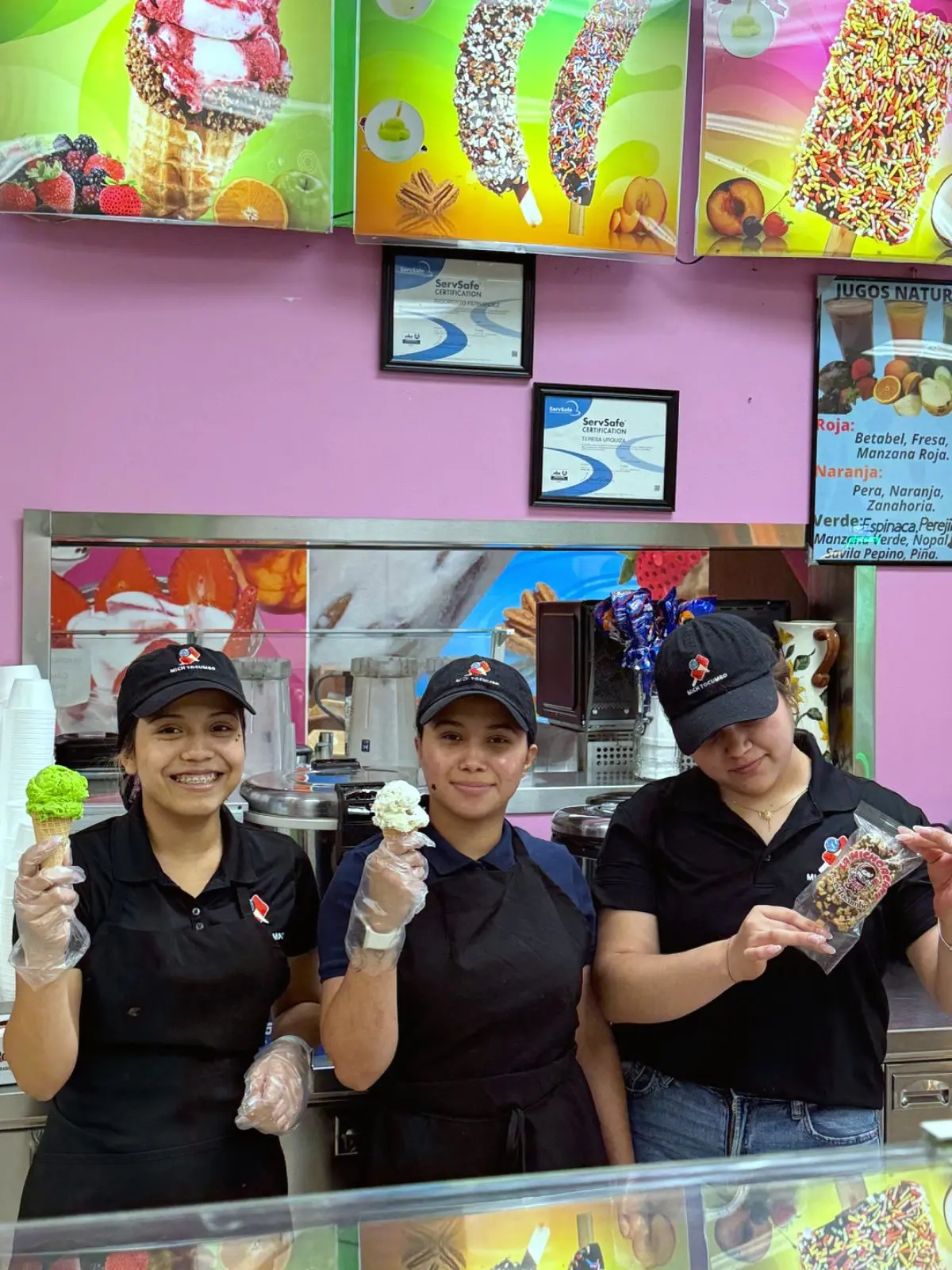 Tres integrantes del equipo sonríen detrás del mostrador con conos y dulces en la mano. Three of the counter crew smiling with cones and treats in hand.
